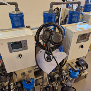 Laboratorieutrustning med blå behållare och digitala pumpar monterade på vita ramar, sammankopplade med svarta slangar och ledningar.