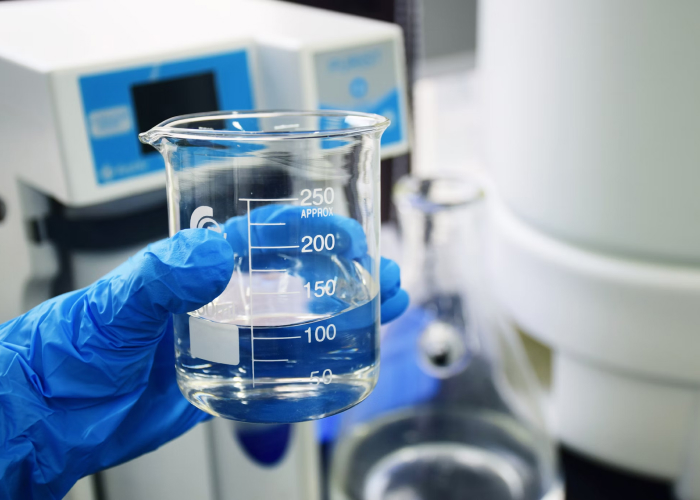 En vetenskapsman som bär en blå handske håller en 250 ml-bägare som innehåller en klar vätska, med laboratorieutrustning i bakgrunden.
