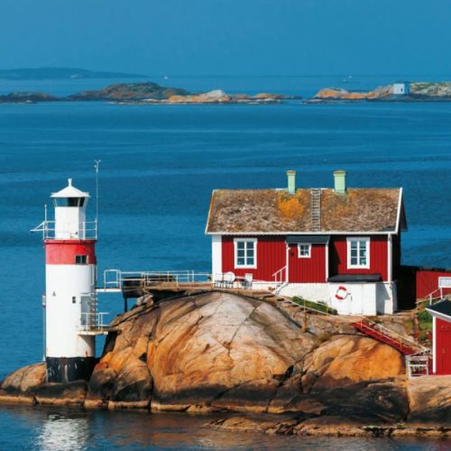 En röd och vit fyr med ett bifogat hus på en stenig kustlinje, med utsikt över havet med avlägsna öar synliga.